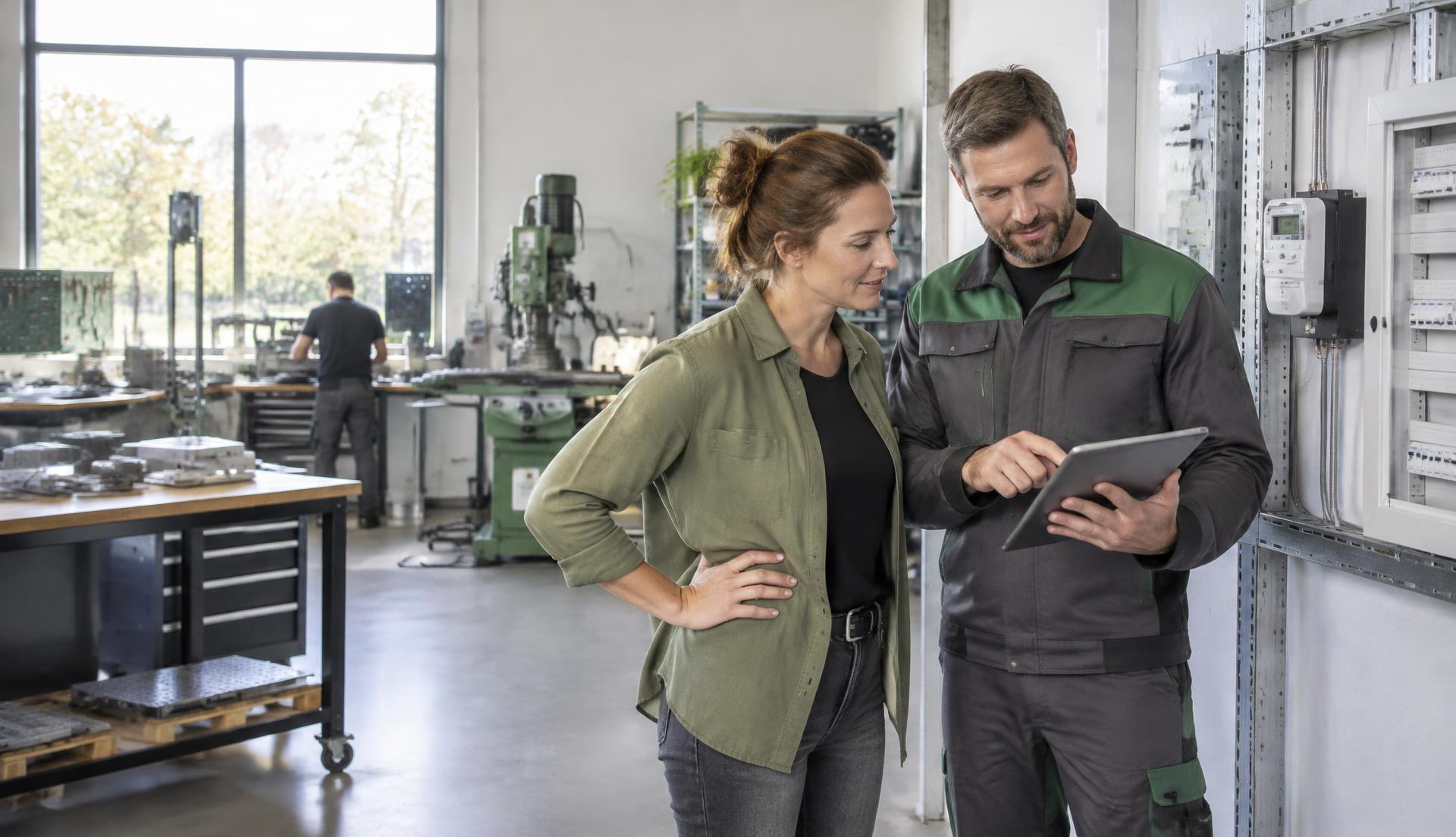 Unternehmerin und Energietechniker prüfen den Stromverbrauch einer Werkstatt am Tablet neben einem Stromzähler